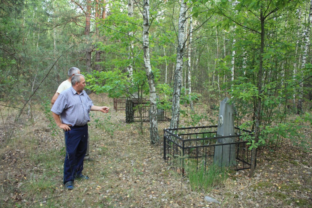 Два чоловіки оглядають могили, окружені металевими огорожами, у лісі з зеленою рослинністю.