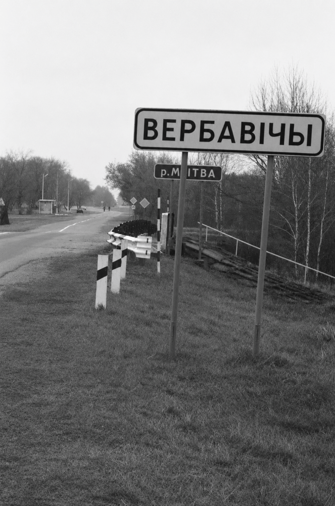 Дорожній знак з написом 'ВЕРБАВИЧІ' та вказівником на річку Мітва, що стоїть на узбіччі дороги в чорнобілому кольорі.