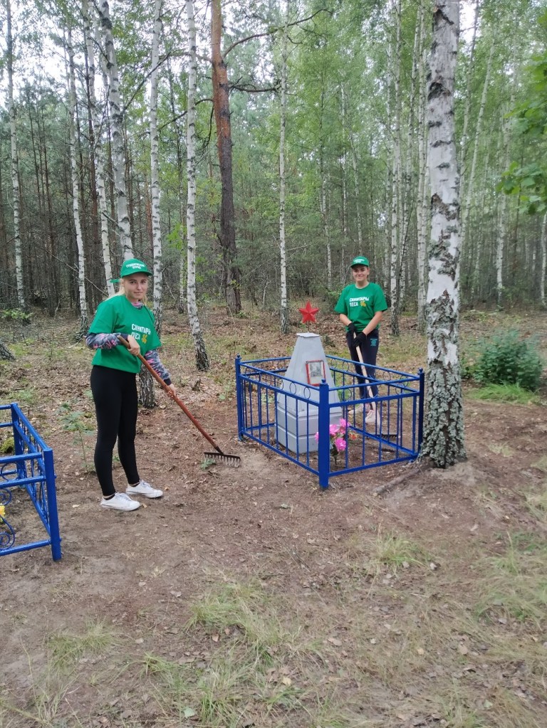 Дві молоді дівчини в зелених футболках прибирають біля пам'ятника в лісі, оточеному деревами. Однією з дівчат у руках граблі, а інша тримає інструмент для роботи в саду.