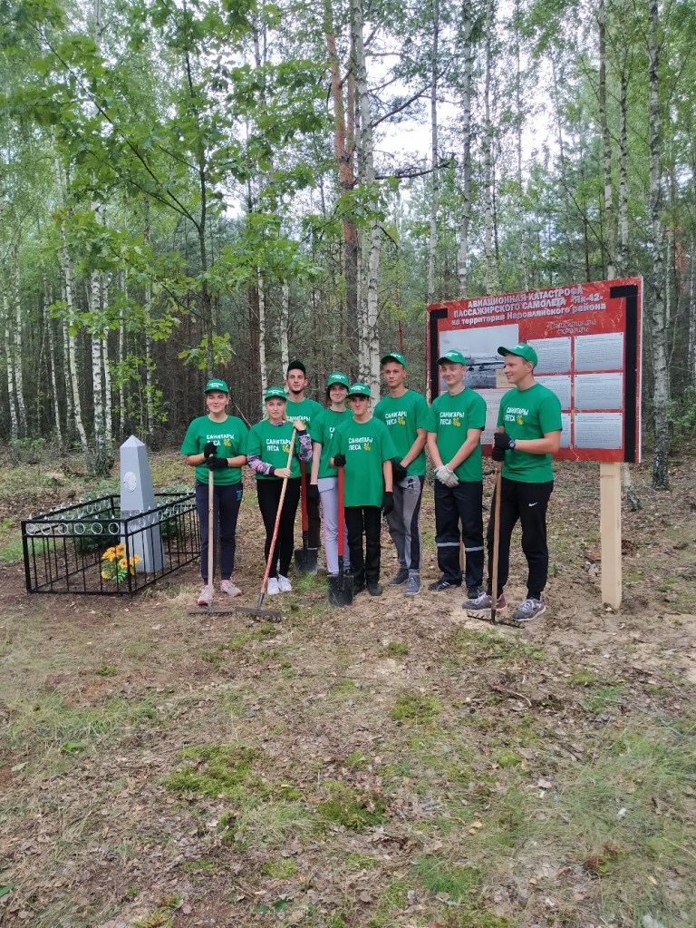 Група молодих людей у зелених футболках позує біля пам'ятника в лісовій місцевості.
