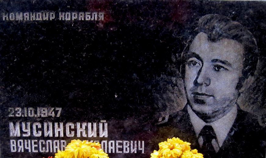 Пам'ятна дошка з портретом Вячеслава Мусинського, командира корабля, з жовтими квітами на підтримку.