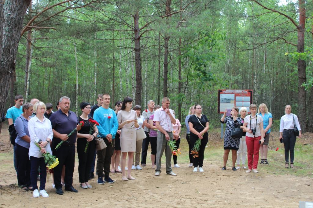 Група людей стоїть у лісі, тримаючи квіти, ймовірно, на memorial service або вшанування пам'яті.