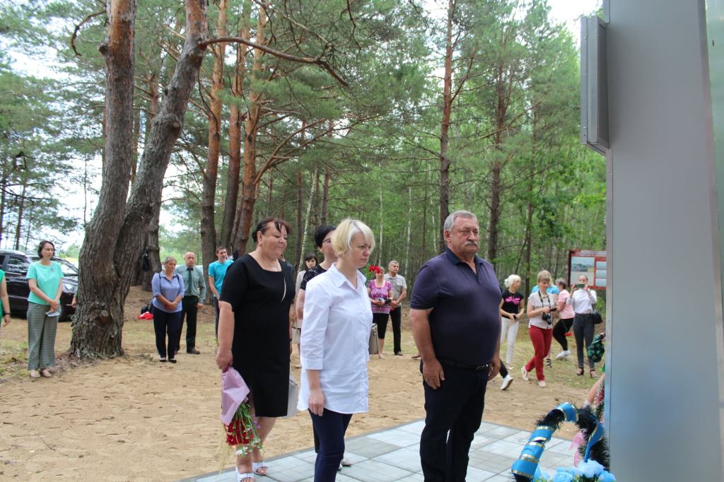 Група людей, що стоїть у лісі, біля пам'ятника, де відбувається вшанування з квітами.