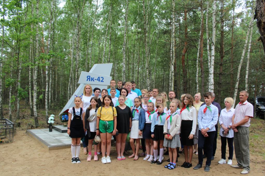 Група людей, включаючи дітей та дорослих, зібралася біля пам'ятника ЯК-42 у лісі.
