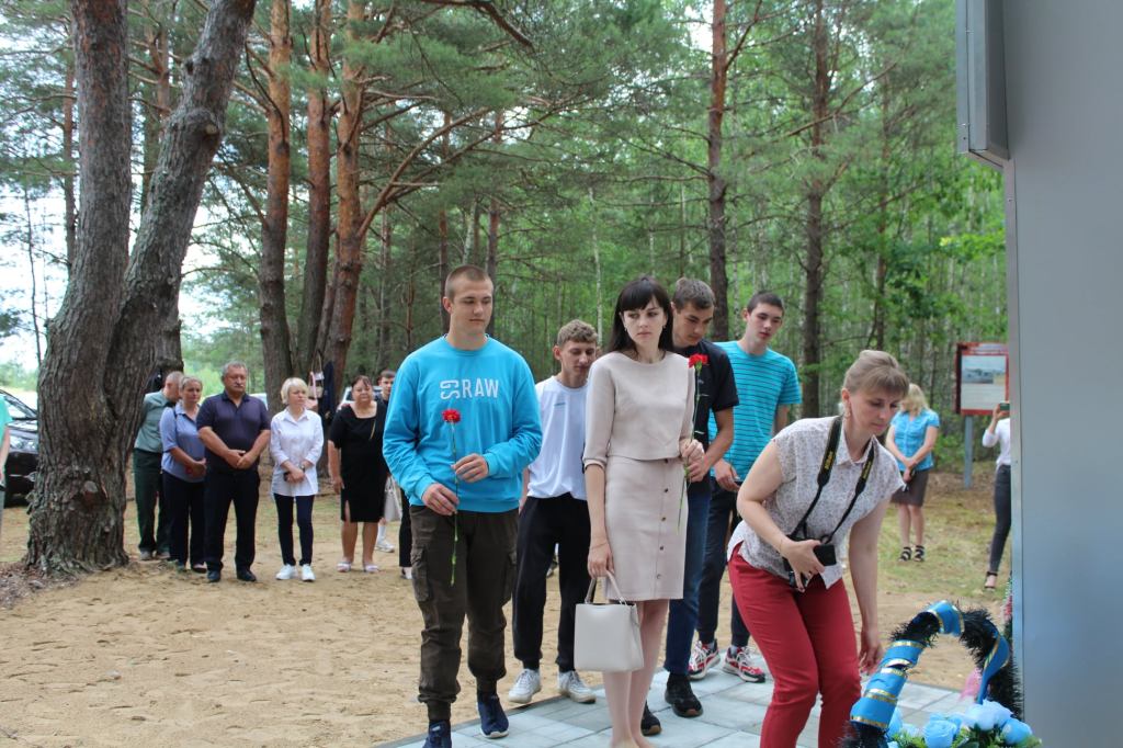 Група людей, серед яких чоловіки і жінки, підходять до пам'ятника в лісі. Деякі тримають квіти, інші виглядають урочисто.